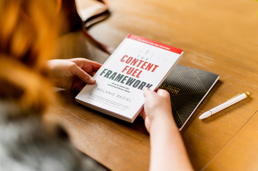 Over the shoulder shot of a red-haired woman reading a book at a cafe, at a wooden table, with a fancy white pen and black notebook nearby.